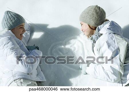 Stock Photograph - Two teen girls lying on snow, looking at heart with initials traced in snow. Fotosearch