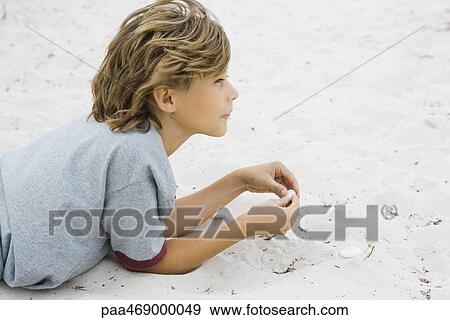 Boy lying on sand, holding pebble, looking away, side view View Large Photo Image Stock Photo - Boy lying on sand, holding pebble, looking away, side view. Fotosearch