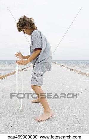 Stock Image - Boy standing on pier, playing with plastic hoop, side view, full length. Fotosearch