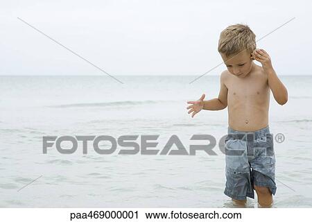 Little boy standing knee deep in ocean, holding up seashell to ear, front view View Large Photo Image Stock Image - Little boy standing knee deep in ocean, holding up seashell to ear, front view. Fotosearch