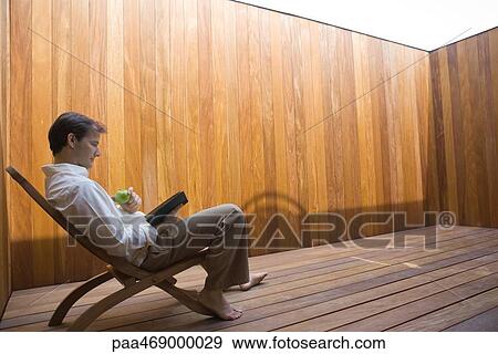 Man sitting in lounge chair, barefoot, reading book and eating apple, full length, side view View Large Photo Image Stock Photo - Man sitting in lounge chair, barefoot, reading book and eating apple, full length, side view . Fotosearch