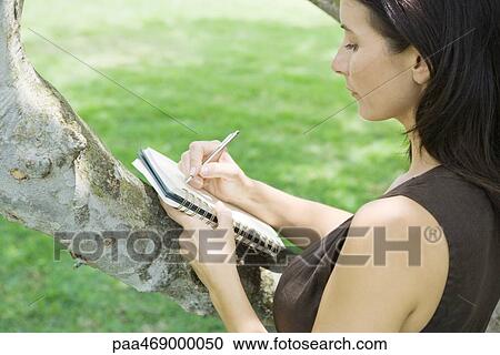 Stock Image - Woman writing in notebook, holding notebook against branch of tree, high angle view. Fotosearch