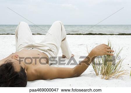 Stock Photo - Young man lying on back on sand, touching dune grass, ocean in background. Fotosearch