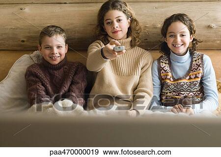 Stock Photo - Teen girl and younger boy and girl watching TV together, front view. Fotosearch