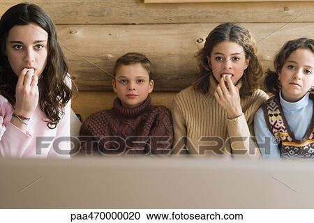 Stock Image - Teen girls and younger boy and girl watching TV together and snacking, front view. Fotosearch