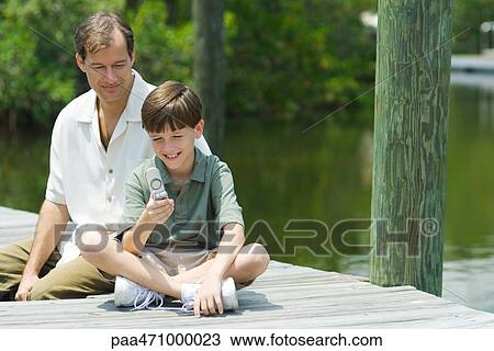 Father and son sitting on dock, looking down at cell phone together View Large Photo Image Stock Image - Father and son sitting on dock, looking down at cell phone together. Fotosearch