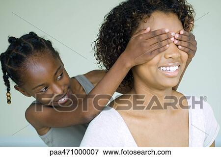 Stock Photo - Girl placing hands over her mother's eyes from behind, both smiling. Fotosearch