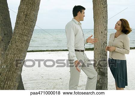 Man and woman standing next to tree trunk at the beach, smiling at each other View Large Photo Image Stock Photo - Man and woman standing next to tree trunk at the beach, smiling at each other. Fotosearch