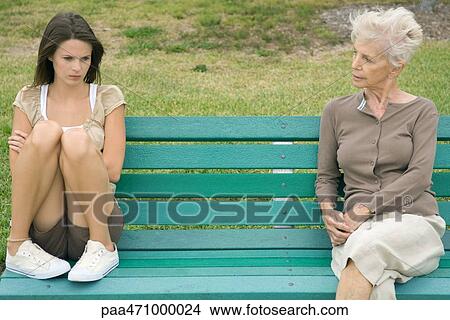 Teenage girl sitting apart from grandmother on bench, arms folded, looking down View Large Photo Image Picture - Teenage girl sitting apart from grandmother on bench, arms folded, looking down. Fotosearch
