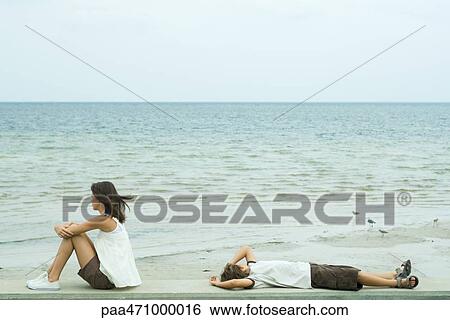 Two young friends looking at ocean, one sitting, the other lying on back View Large Photo Image Stock Photograph - Two young friends looking at ocean, one sitting, the other lying on back. Fotosearch