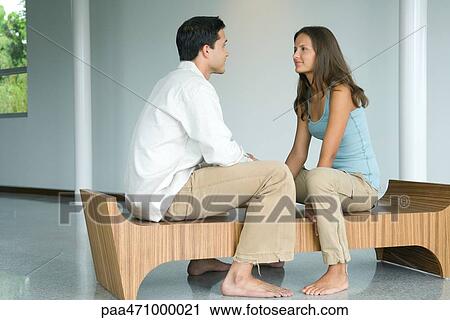 Stock Image - Young couple sitting face to face on bench, smiling at each other. Fotosearch