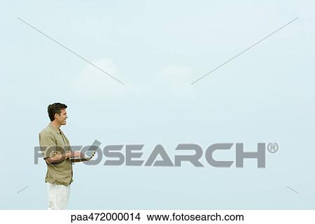Man standing outdoors holding laptop computer, looking away View Large Photo Image Picture - Man standing outdoors holding laptop computer, looking away. Fotosearch