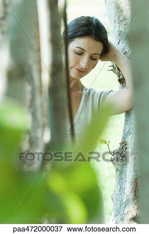 Woman leaning against tree trunk, looking down, view through foliage View Large Photo Image Stock Photo - Woman leaning against tree trunk, looking down, view through foliage. Fotosearch