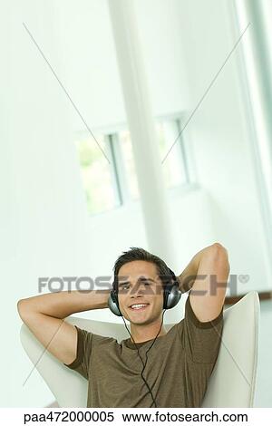 Young man listening to headphones with hands behind head, smiling at camera View Large Photo Image Stock Photography - Young man listening to headphones with hands behind head, smiling at camera. Fotosearch