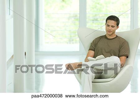 Stock Photo - Young man sitting in chair reading book. Fotosearch
