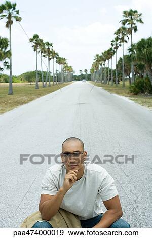 Man sitting in the middle of road with hand under chin, looking away, car in the distance View Large Photo Image Stock Photo - Man sitting in the middle of road with hand under chin, looking away, car in the distance. Fotosearch
