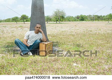 Stock Photo - Man sitting on ground leaning against suitcase, looking at camera. Fotosearch