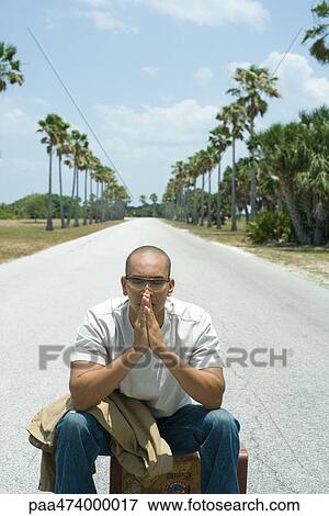 Stock Photo - Man sitting on suitcase in the middle of road, eyes closed, hands clasped in front of face. Fotosearch
