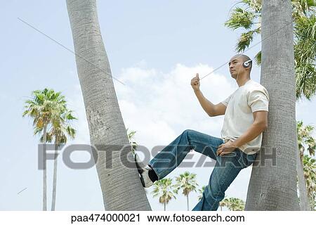 Man wearing headphones leaning against tree with thumb raised, looking away View Large Photo Image Stock Image - Man wearing headphones leaning against tree with thumb raised, looking away. Fotosearch