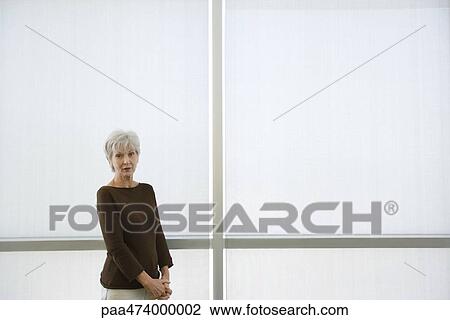 Stock Image - Senior woman standing with clasped hands, looking at camera. Fotosearch