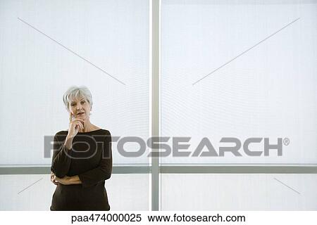 Stock Photography - Senior woman standing with hand under chin, looking at camera. Fotosearch