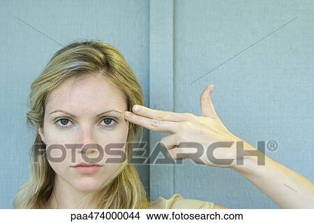 Picture - Woman holding fingers against temple in gun gesture, looking at camera. Fotosearch