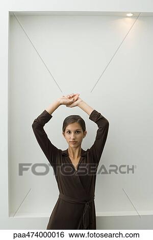 Stock Photograph - Young female leaning against wall with arms raised, looking at camera. Fotosearch