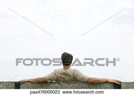 Man sitting on bench with arms outstretched, looking at sky, rear view View Large Photo Image Stock Image - Man sitting on bench with arms outstretched, looking at sky, rear view. Fotosearch