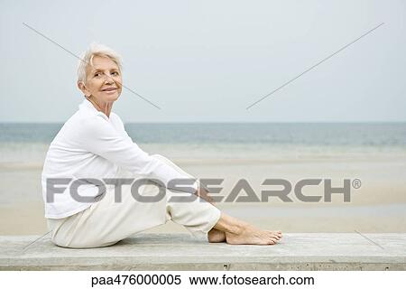 Stock Photography - Senior woman sitting at the beach, looking up and smiling. Fotosearch