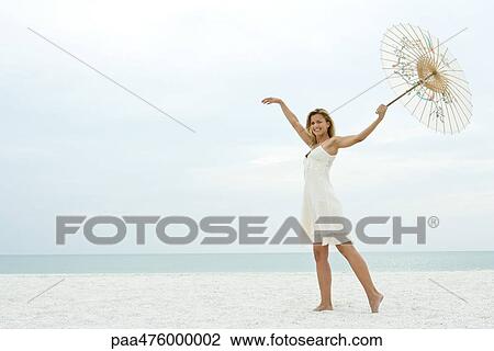 Stock Image - Woman standing on beach with arms raised, holding parasol, smiling. Fotosearch