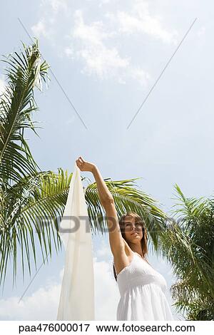 Stock Photo - Woman standing under palm tree holding scarf, low angle view. Fotosearch