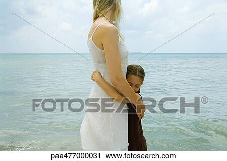 Stock Image - Mother and daughter standing by water, hugging, ocean horizon in background. Fotosearch