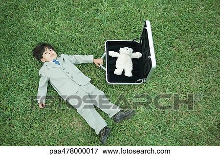 Stock Photo - Little boy in full suit, lying on grass, next to open briefcase containing teddy bear. Fotosearch