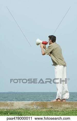 Stock Image - Man using megaphone, facing ocean horizon, full length. Fotosearch