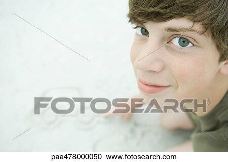 Stock Image - Teen boy drawing car in sand, looking up at camera. Fotosearch