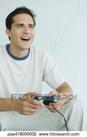Stock Image - Young man playing video game, looking out of frame, smiling. Fotosearch