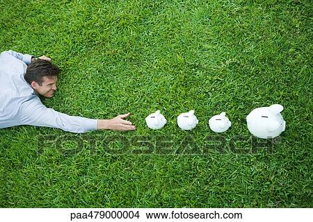 Picture - Man lying on grass, reaching for line of piggy banks. Fotosearch
