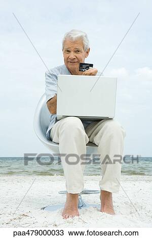 Senior man making credit card purchase online, holding up credit card, sitting in chair on beach View Large Photo Image Stock Image - Senior man making credit card purchase online, holding up credit card, sitting in chair on beach. Fotosearch