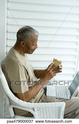 Stock Photo - Senior man making credit card purchase online, looking at number on credit card. Fotosearch