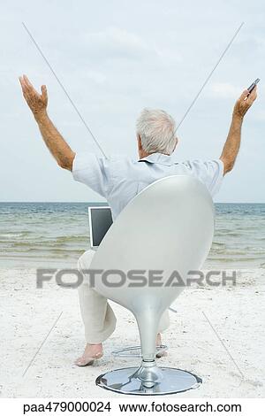 Senior man sitting in chair on beach, using laptop, arms in air, rear view View Large Photo Image Picture - Senior man sitting in chair on beach, using laptop, arms in air, rear view. Fotosearch