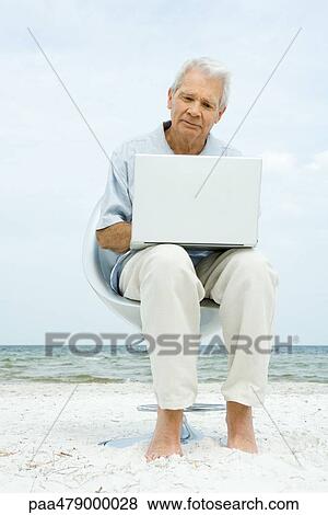 Senior man sitting in chair on beach, using laptop View Large Photo Image Stock Photo - Senior man sitting in chair on beach, using laptop. Fotosearch