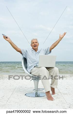 Stock Image - Senior man sitting on beach with laptop on lap, arms raised, shouting. Fotosearch