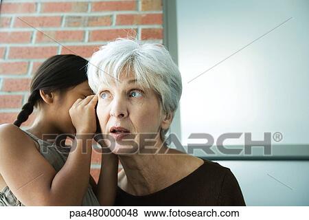 Stock Photo - Girl whispering in grandmother's ear, close-up. Fotosearch