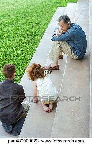 Stock Image - Grandfather and two grandchildren playing with toy cars together on stairs, high angle view. Fotosearch