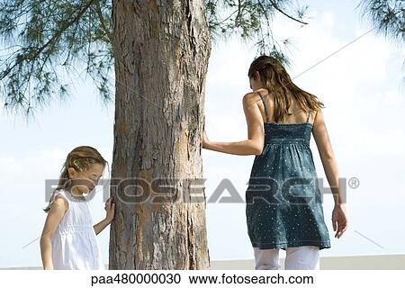 Stock Image - Two sisters walking around tree, touching trunk, low angle view. Fotosearch
