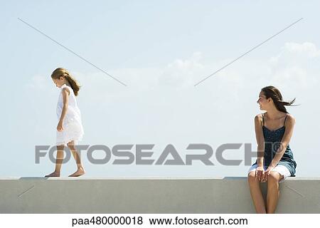 Stock Photo - Young woman sitting outdoors, watching younger sister walk across ledge. Fotosearch