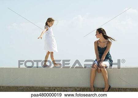 Stock Photo - Young woman sitting outdoors, watching younger sister walk across ledge. Fotosearch