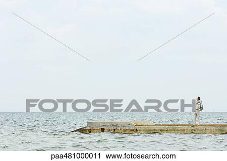 Stock Image - Man standing on pier, looking up, in the distance. Fotosearch