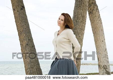 Stock Photo - Woman leaning against tree trunk, looking away. Fotosearch