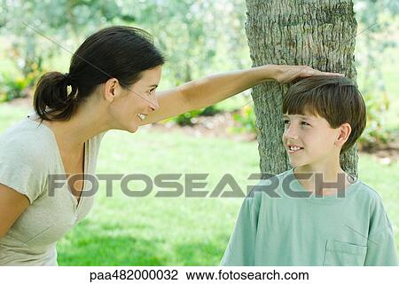 Stock Image - Boy leaning against tree trunk, his mother placing her hand on his head, both smiling. Fotosearch